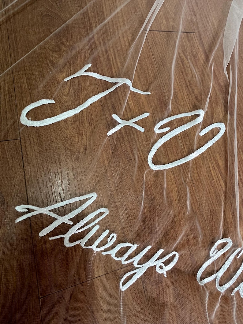 May include: A sheer white bridal veil with embroidered white text. The text includes the initials "J x C" and the words "Always & Forever". The veil is draped over a wooden surface.