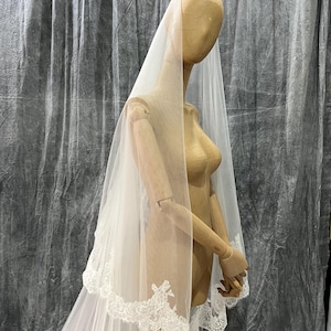May include: A sheer white bridal veil with a delicate lace trim. The veil is draped over a wooden mannequin, showcasing its flowing design. The lace trim adds a touch of elegance to this wedding accessory.