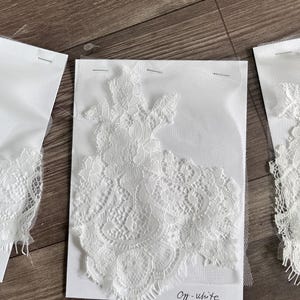 May include: Off-white lace fabric samples pinned to white cards. The lace features intricate floral patterns and delicate edges. The cards are labeled "Off-white" in handwritten script. The samples are displayed on a wooden surface.