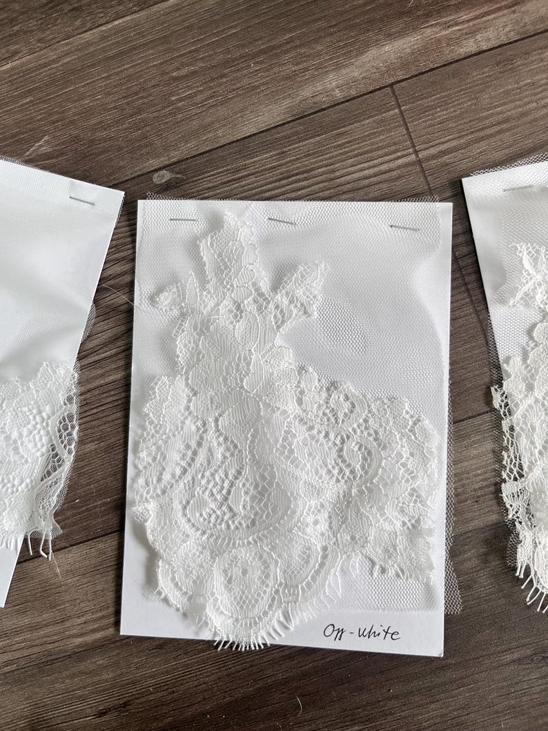 May include: Off-white lace fabric samples pinned to white cards. The lace features intricate floral patterns and delicate edges. The cards are labeled "Off-white" in handwritten script. The samples are displayed on a wooden surface.