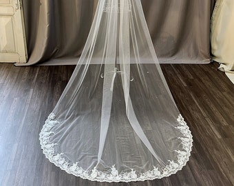 May include: A white bridal veil with a delicate lace trim and scattered pearl embellishments. The veil is long and flowing, with a scalloped edge.