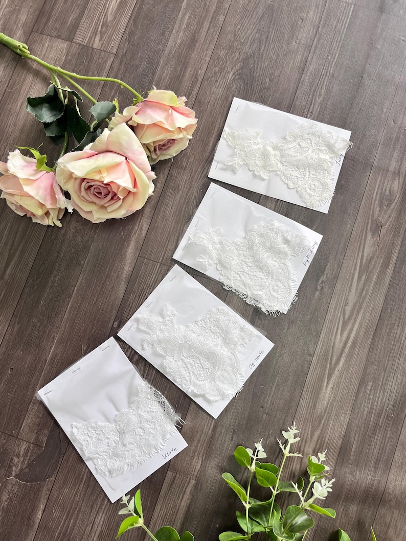May include: Four samples of white lace fabric on white cards, labeled "White", "Off-white", and "Light Ivory". The lace has intricate floral patterns. The image also includes pink roses and greenery, all arranged on a wooden surface.