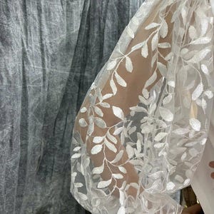 May include: White sheer sleeve with an embroidered leaf pattern. The sleeve is part of a white garment, with a gathered design at the shoulder and wrist. The background is a textured gray fabric.