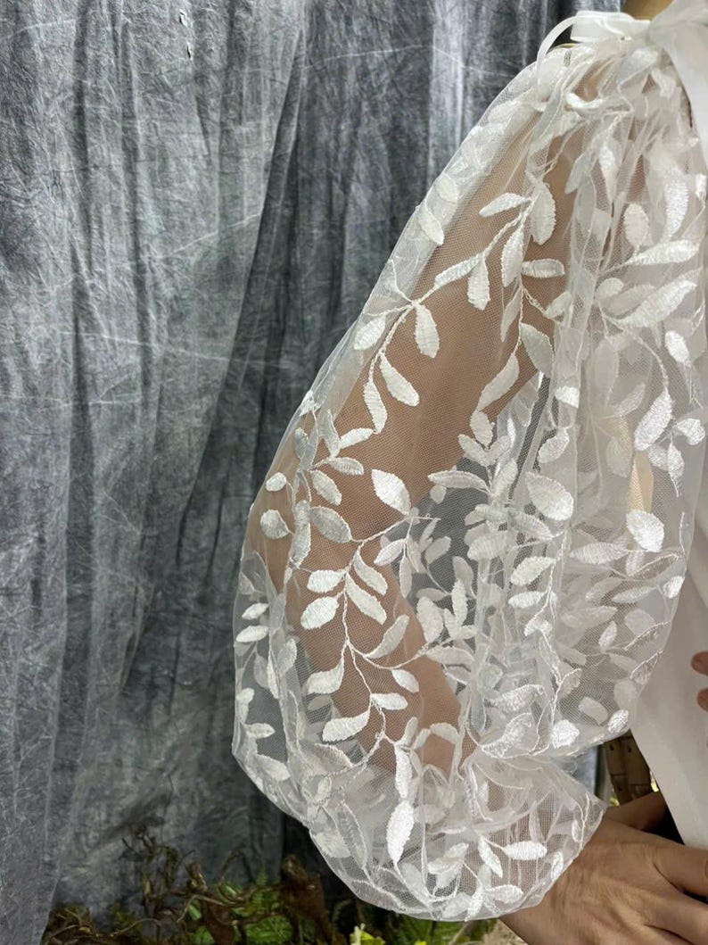 May include: White sheer sleeve with an embroidered leaf pattern. The sleeve is part of a white garment, with a gathered design at the shoulder and wrist. The background is a textured gray fabric.