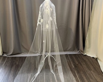 May include: A long, sheer white bridal veil with delicate floral lace detailing along the edges. The veil is adorned with small, scattered pearls and is displayed on a wire mannequin. The veil is a classic wedding accessory.