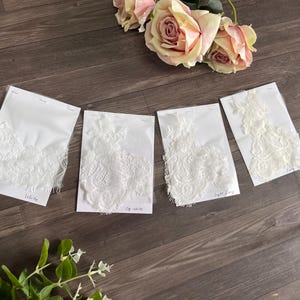 May include: Four white lace fabric samples on white cards, labeled "White", "Off White", and "Light Ivory". The samples are arranged on a wooden surface, with pink roses in the background.