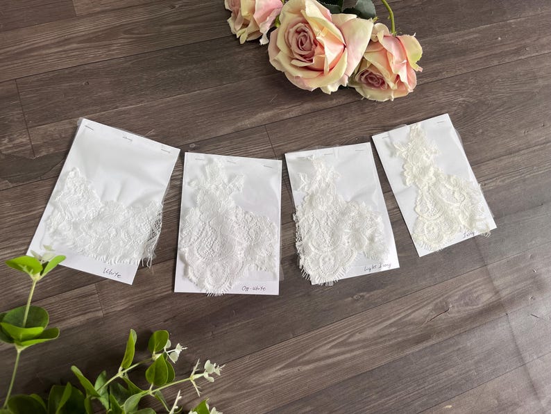 May include: Four white lace fabric samples on white cards, labeled "White", "Off White", and "Light Ivory". The samples are arranged on a wooden surface, with pink roses in the background.