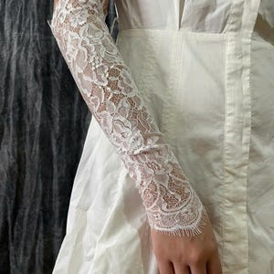 May include: A white lace sleeve accessory featuring an intricate floral pattern with scalloped edges. The delicate fabric extends from the upper arm to the wrist, showcasing a detailed mesh design that is worn over a plain white sleeveless garment.