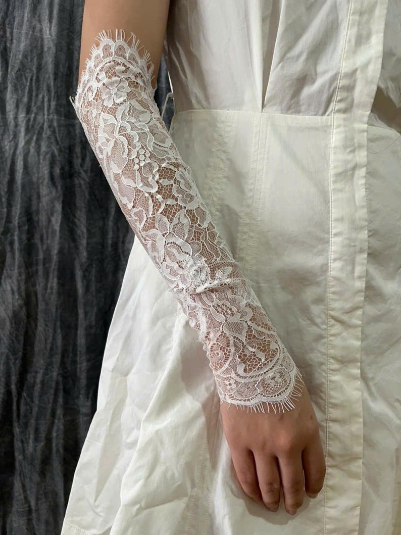 May include: A white lace sleeve accessory featuring an intricate floral pattern with scalloped edges. The delicate fabric extends from the upper arm to the wrist, showcasing a detailed mesh design that is worn over a plain white sleeveless garment.