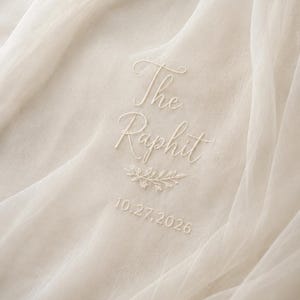 May include: Ivory fabric with embroidered text reading "The Raphit" above a sprig of leaves and the date "10.27.2026". The fabric is draped, creating soft folds and shadows. The embroidery is in a matching ivory color.
