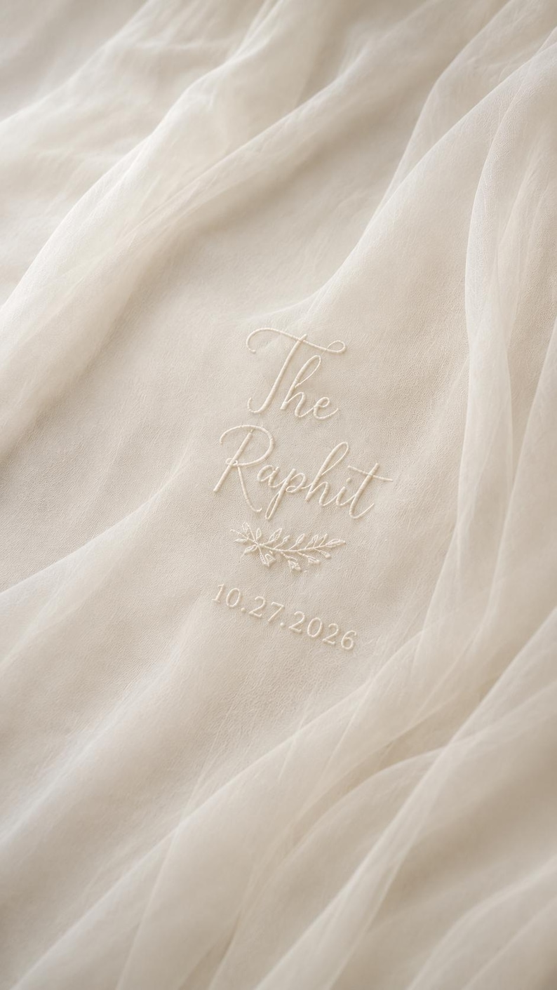 May include: Ivory fabric with embroidered text reading "The Raphit" above a sprig of leaves and the date "10.27.2026". The fabric is draped, creating soft folds and shadows. The embroidery is in a matching ivory color.