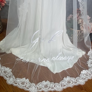 May include: A white bridal veil with delicate lace trim and embroidered initials and the words "Forever always". The veil is sheer and drapes over a white dress, creating an elegant look for a wedding.