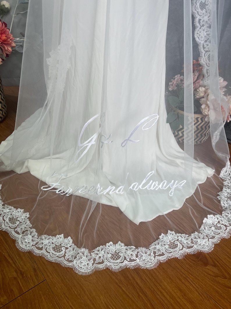 May include: A white bridal veil with delicate lace trim and embroidered initials and the words "Forever always". The veil is sheer and drapes over a white dress, creating an elegant look for a wedding.