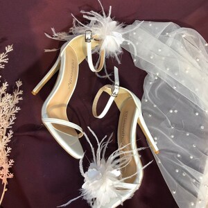 May include: A pair of white satin high heel sandals with delicate feather and flower embellishments. The sandals have a thin ankle strap and the brand name "Delorenzi" is visible on the insole.