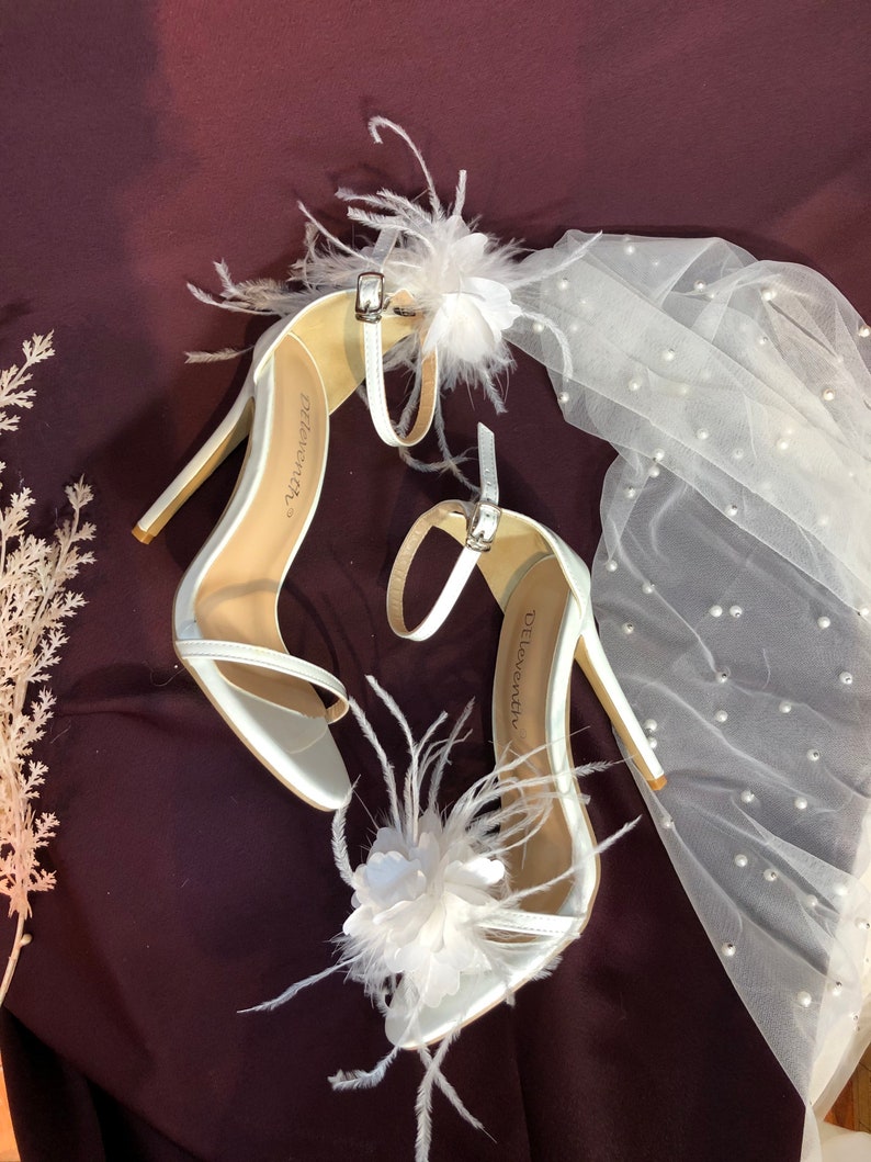 May include: A pair of white satin high heel sandals with delicate feather and flower embellishments. The sandals have a thin ankle strap and the brand name "Delorenzi" is visible on the insole.