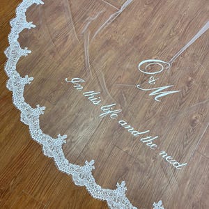 May include: A sheer white wedding veil with a delicate lace trim. The veil features the embroidered text "In this life and the next" along with the initials "O & M". The lace trim is white and scalloped.