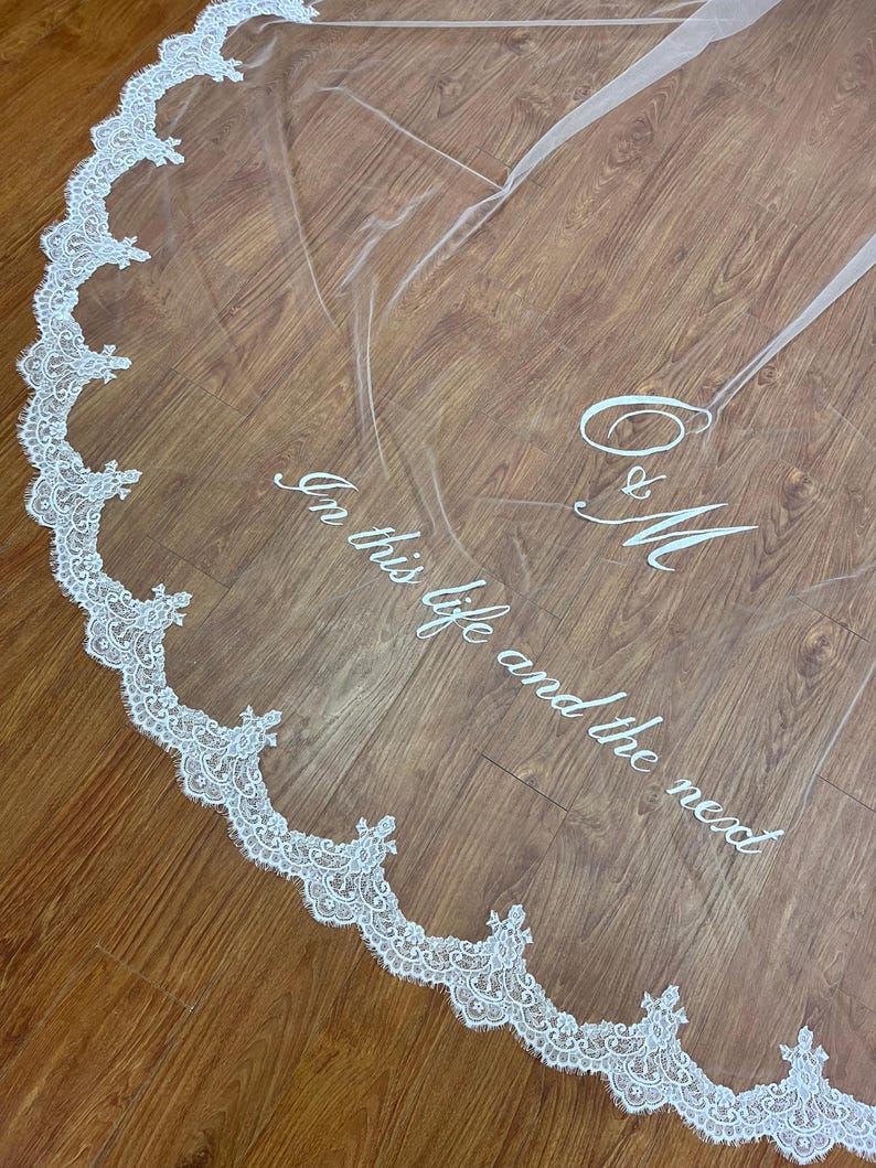 May include: A sheer white wedding veil with a delicate lace trim. The veil features the embroidered text "In this life and the next" along with the initials "O & M". The lace trim is white and scalloped.