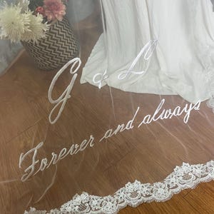 May include: A sheer white wedding veil with embroidered script that reads "G & L Forever and always." The veil has a delicate lace trim along the bottom edge. A white wedding dress is visible in the background.