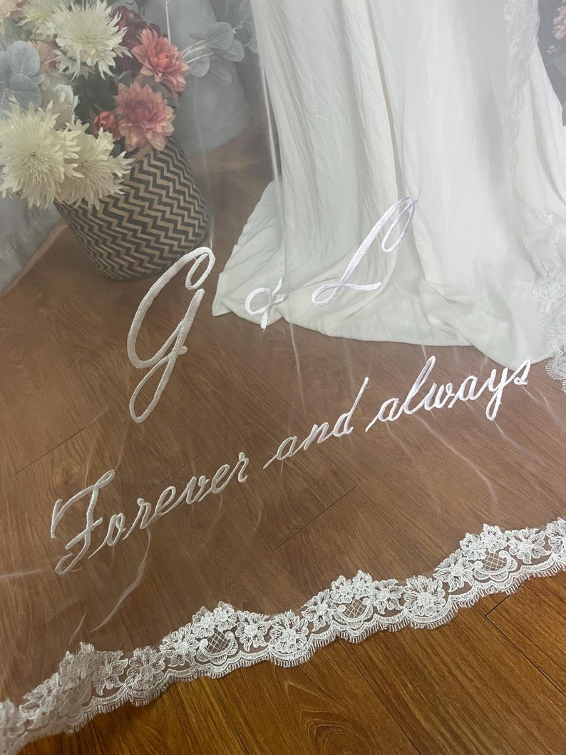 May include: A sheer white wedding veil with embroidered script that reads "G & L Forever and always." The veil has a delicate lace trim along the bottom edge. A white wedding dress is visible in the background.