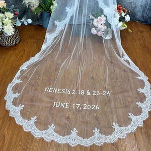 May include: A white wedding veil with a delicate lace trim. The veil features the text "GENESIS 2:18 & 23-24" and "JUNE 17, 2026" in a simple font. The veil is draped over a wooden surface.