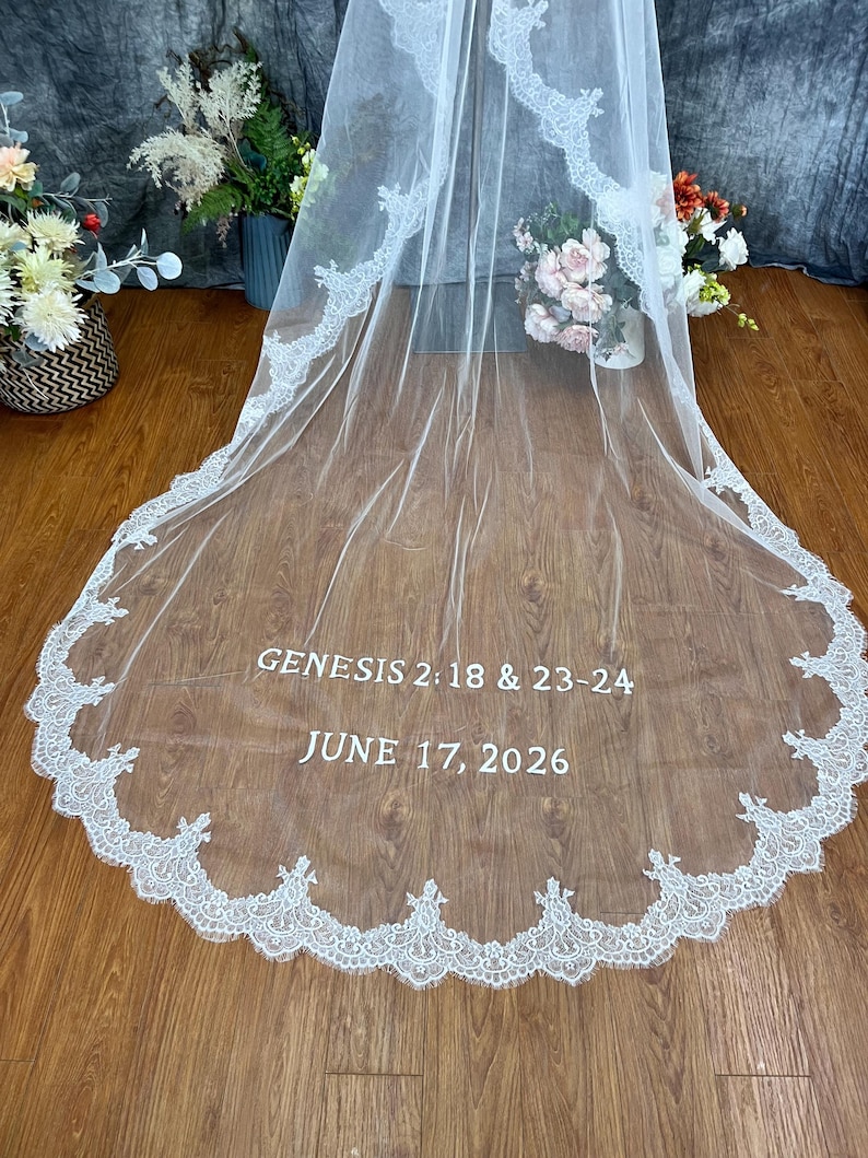 May include: A white wedding veil with a delicate lace trim. The veil features the text "GENESIS 2:18 & 23-24" and "JUNE 17, 2026" in a simple font. The veil is draped over a wooden surface.