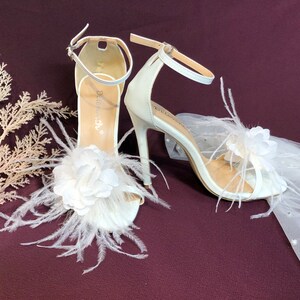 May include: White bridal sandals with a feather and flower embellishment. The sandals have a high heel and an ankle strap.