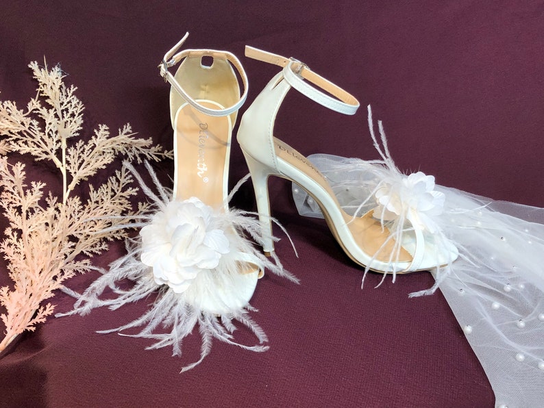 May include: White bridal sandals with a feather and flower embellishment. The sandals have a high heel and an ankle strap.