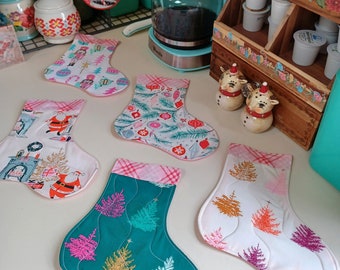 Op de afbeelding: Een verzameling kerstman-vormige onderzetters met diverse feestelijke patronen. Ontwerpen omvatten kerstbomen, ornamenten en de kerstman. De onderzetters zijn gemaakt van stof en hebben gewatteerde details. Het kleurenpalet omvat rood, groen, roze en wit.