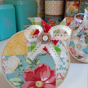 May include: A round, white-framed embroidery hoop with a patchwork design featuring floral patterns in shades of blue, red, yellow, and green. A white lace bow with a green and white checkered ribbon and a pearl button embellishment is attached to the hoop.