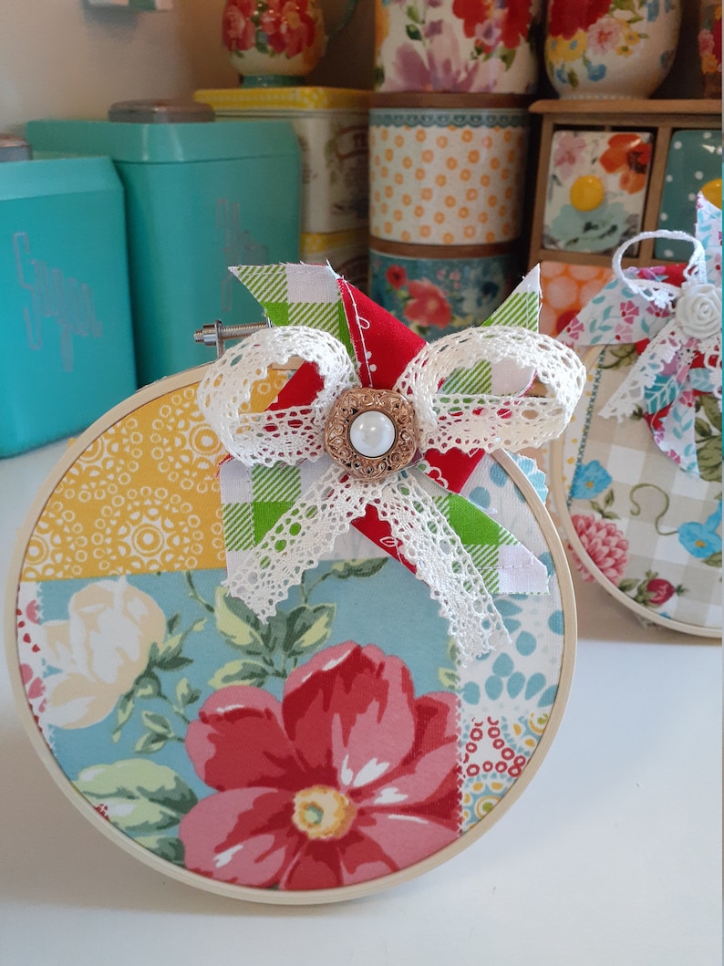 May include: A round, white-framed embroidery hoop with a patchwork design featuring floral patterns in shades of blue, red, yellow, and green. A white lace bow with a green and white checkered ribbon and a pearl button embellishment is attached to the hoop.