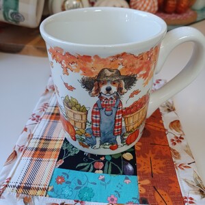 May include: A white ceramic mug with a fall-themed design featuring a dog wearing overalls and holding a basket of apples. The mug is sitting on a patchwork fabric coaster with a plaid pattern.