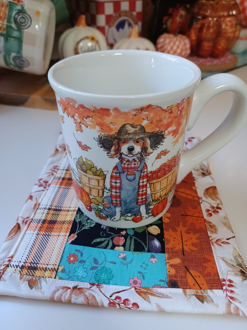 May include: A white ceramic mug with a fall-themed design featuring a dog wearing overalls and holding a basket of apples. The mug is sitting on a patchwork fabric coaster with a plaid pattern.