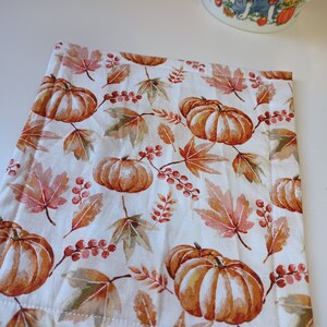 May include: A white fabric coaster with a fall-themed design featuring orange pumpkins, brown leaves, and red berries.