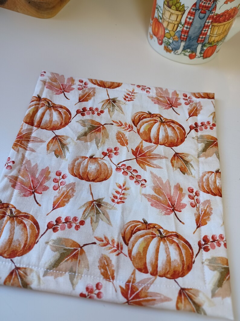May include: A white fabric coaster with a fall-themed design featuring orange pumpkins, brown leaves, and red berries.