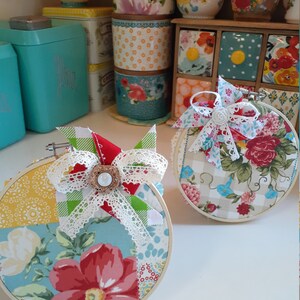 May include: Two round embroidery hoops with fabric featuring a patchwork design of floral patterns, gingham, and polka dots. The fabric is in shades of red, blue, green, yellow, and white. Each hoop has a white lace bow with a pearl button in the center.