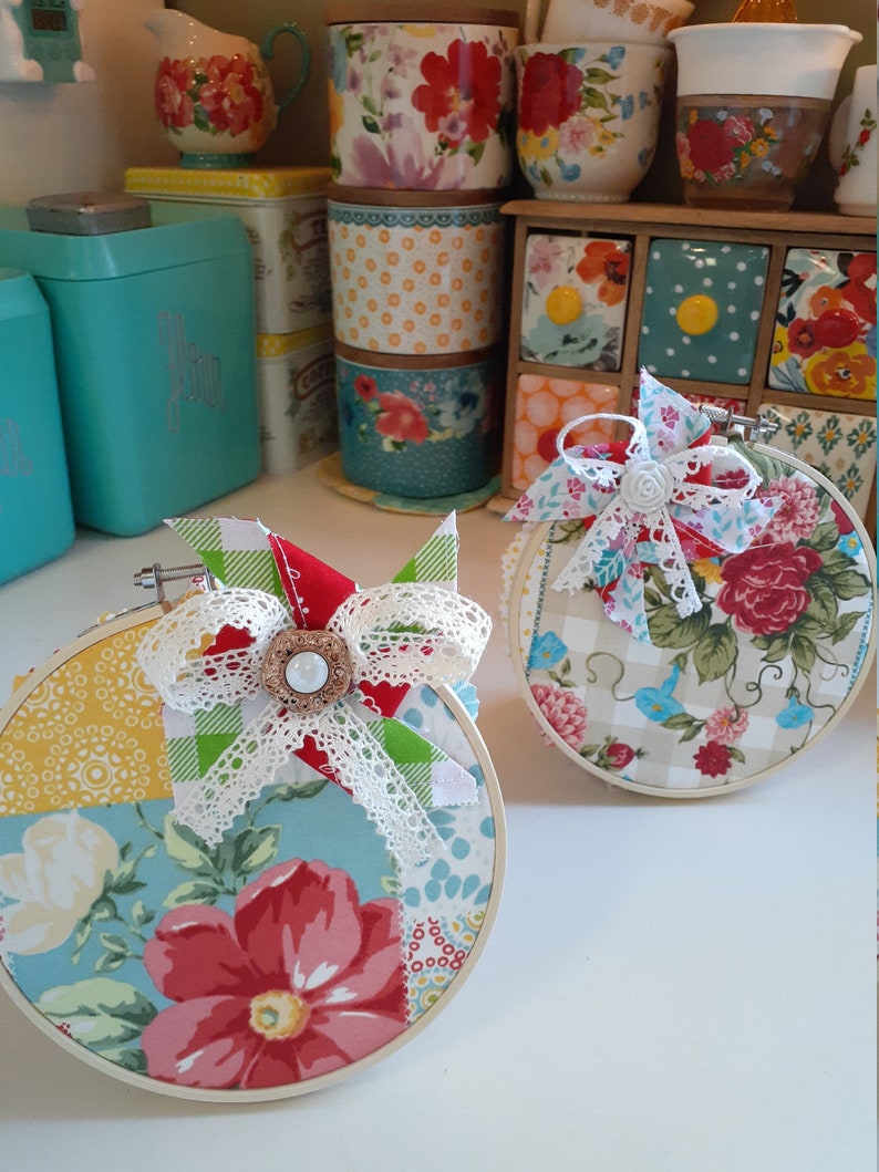 May include: Two round embroidery hoops with fabric featuring a patchwork design of floral patterns, gingham, and polka dots. The fabric is in shades of red, blue, green, yellow, and white. Each hoop has a white lace bow with a pearl button in the center.