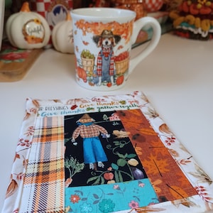 May include: A white ceramic mug with a fall-themed design featuring a dog wearing a blue and white plaid shirt and a brown hat. The mug is sitting on a patchwork fabric coaster with a scarecrow and the text "Give thanks & gather together" in brown lettering.