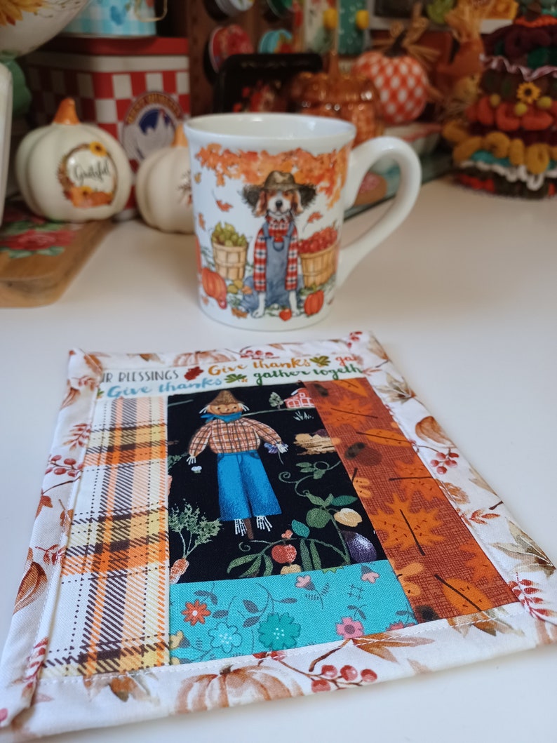 May include: A white ceramic mug with a fall-themed design featuring a dog wearing a blue and white plaid shirt and a brown hat. The mug is sitting on a patchwork fabric coaster with a scarecrow and the text "Give thanks & gather together" in brown lettering.