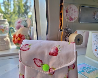 Puede incluir: Una bolsa de tela rosa con cierre de solapa y un botón verde. La bolsa está decorada con un patrón de lunares blancos y una impresión de fresas.