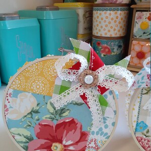May include: Two round embroidery hoops with a patchwork design of floral and geometric patterns in shades of red, yellow, blue, and green. Each hoop has a white lace bow with a pearl button in the center.