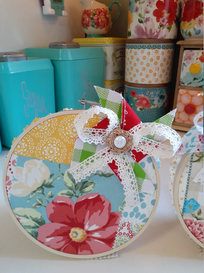 May include: Two round embroidery hoops with a patchwork design of floral and geometric patterns in shades of red, yellow, blue, and green. Each hoop has a white lace bow with a pearl button in the center.