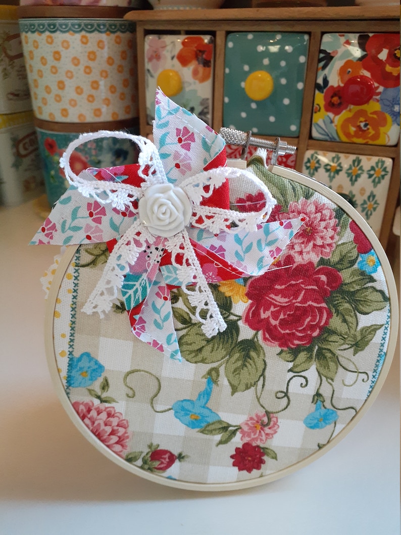 May include: A white embroidery hoop with a floral fabric design. The fabric features a large red rose, smaller pink and blue flowers, and green leaves. A white lace bow with a white fabric flower is attached to the hoop.