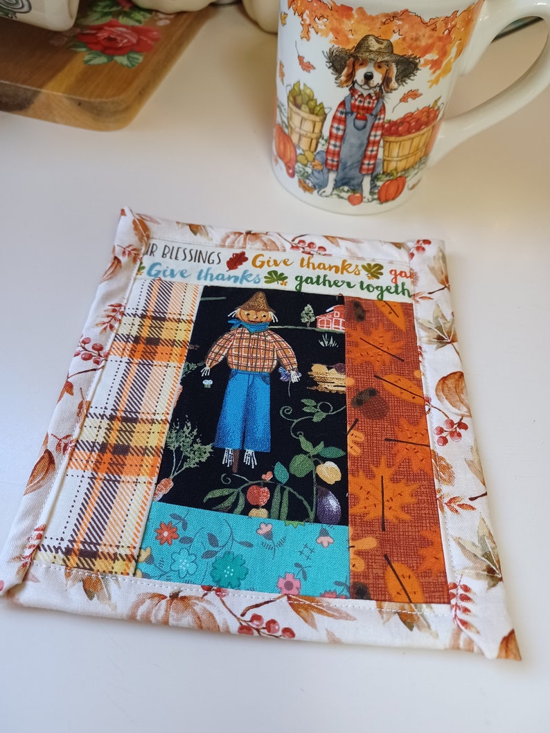 May include: A colorful fabric coaster with a patchwork design featuring a scarecrow, plaid, floral, and fall leaf patterns. The coaster has a white border and the text "Give thanks gather together" is printed on it.
