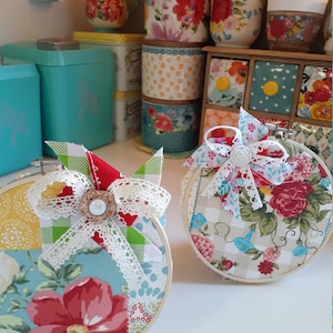 May include: Two round embroidery hoops with floral fabric designs. One hoop has a red, white, and green patchwork design with a white lace bow and a pearl button. The other hoop has a beige and blue floral design with a white lace bow and a white pearl button.