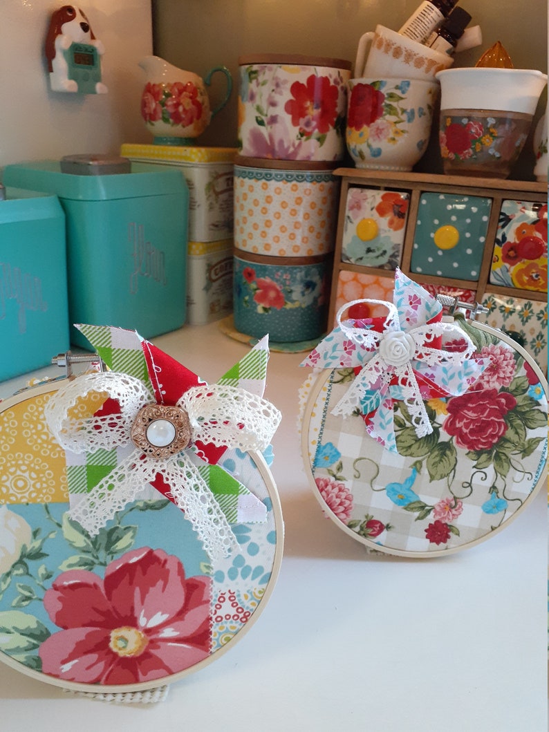 May include: Two round embroidery hoops with floral fabric designs. One hoop has a red, white, and green patchwork design with a white lace bow and a pearl button. The other hoop has a beige and blue floral design with a white lace bow and a white pearl button.