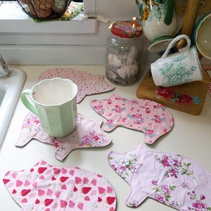 May include: A collection of pink pig-shaped fabric coasters with floral and strawberry patterns. A green mug sits on one coaster. Other kitchen items include a jar, mugs, and a wire basket with eggs.