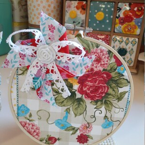 May include: A round embroidery hoop with a floral fabric design. The fabric is a white and beige gingham with a floral pattern in shades of red, pink, blue, and green. A white lace bow with a white fabric flower is attached to the top of the hoop.
