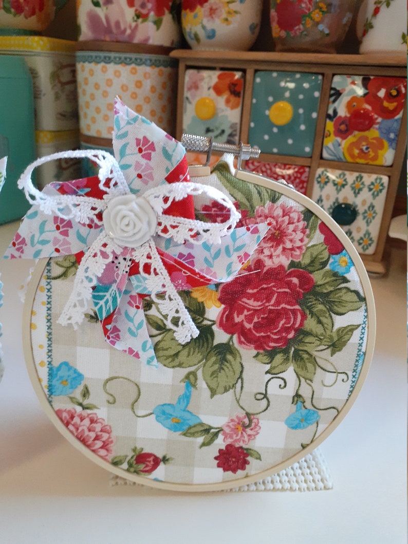 May include: A round embroidery hoop with a floral fabric design. The fabric is a white and beige gingham with a floral pattern in shades of red, pink, blue, and green. A white lace bow with a white fabric flower is attached to the top of the hoop.