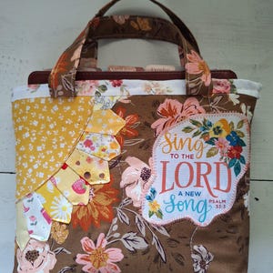 May include: A brown fabric tote bag with a floral pattern, featuring a yellow sun-shaped applique on the left and a white patch on the right with the text Sing TO THE LORD A NEW Song PSALM 33:3 surrounded by colorful floral illustrations.