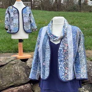 May include: Two quilted jackets, one for a child and one for an adult, on mannequins. The jackets have a patchwork design with floral patterns in blue, grey, and white. Both have navy blue trim. The adult jacket is styled with a navy top and a matching scarf.